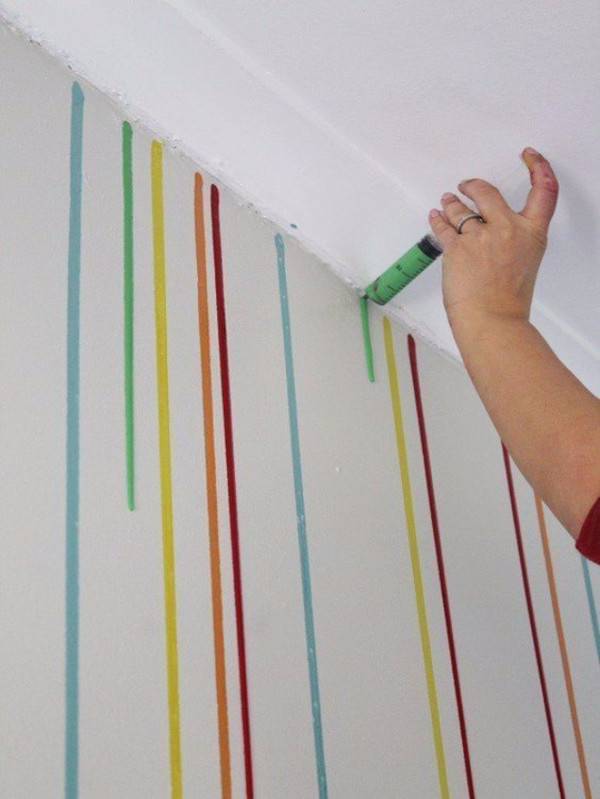A person holding a syringe full of paint. They are squeezing it onto the top of the wall and letting it drip down.